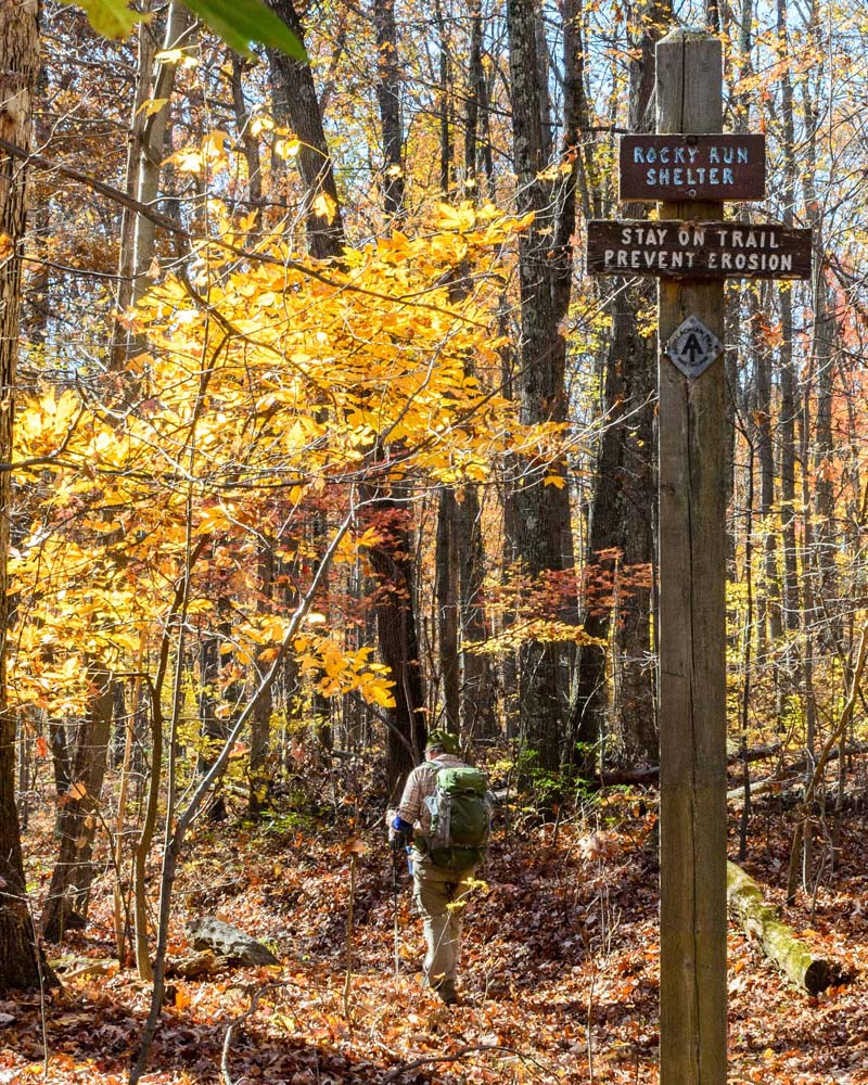 Side trail with sign indicating that it leads to the Rocky Run Shelter.  A hiker is visible going down that trail