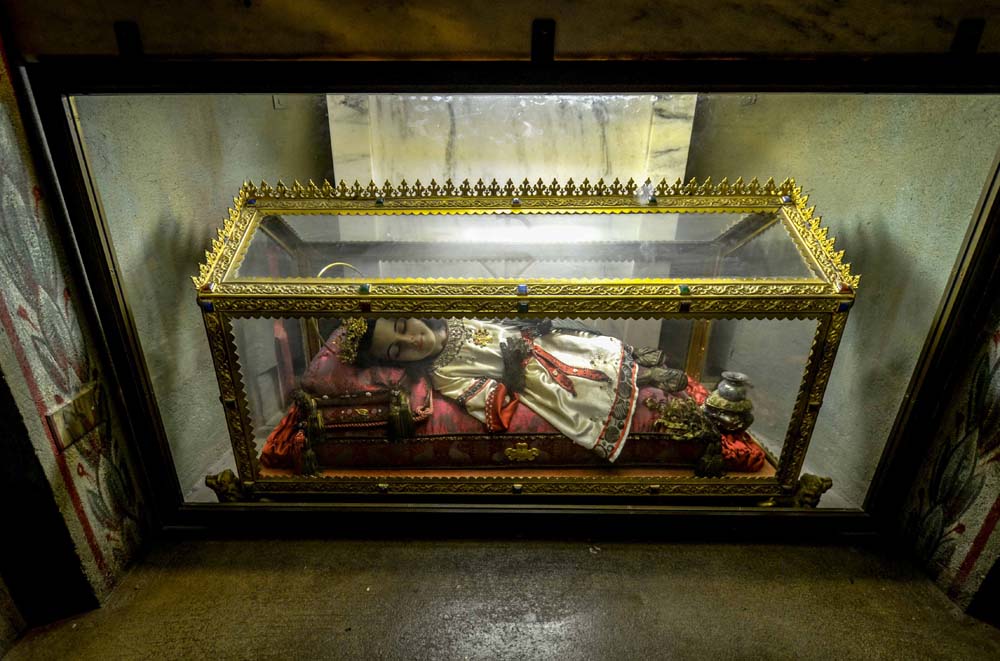 Mummified corpse of Saint Innocent wearing ornate embroidered dres, crown, with jar at feet with saints blood