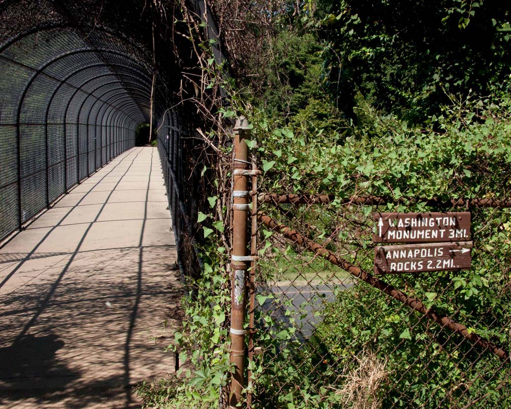 Foot bridge with chain-link fencing over the top of it to prevent people falling off.  The bridge crosses over a four lane highway.  A sign indicates trail directions north and south